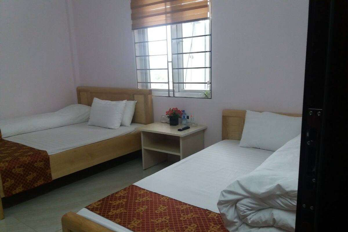A hotel room with two single beds, a small bedside table with a lamp, and a window with blinds.