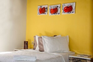 Een bed met witte beddengoed, een houten nachtkastje en een gele muur met drie rode appel schilderijen.