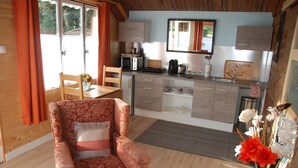 Une pièce chaleureuse avec un plancher en bois, un fauteuil à motifs floraux, une table à manger avec des chaises, une petite cuisine équipée d’un four à micro-ondes et une grande fenêtre avec des rideaux.