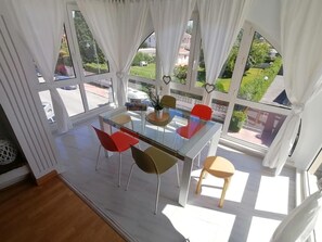 Un'area pranzo con tavolo e sedie in vetro, ampie finestre con tende e vista sul verde.