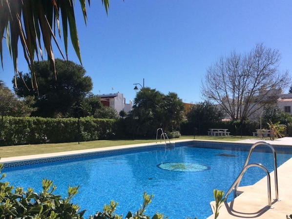 Una piscina rettangolare con acqua cristallina, circondata da vegetazione curata e alcuni edifici sullo sfondo.