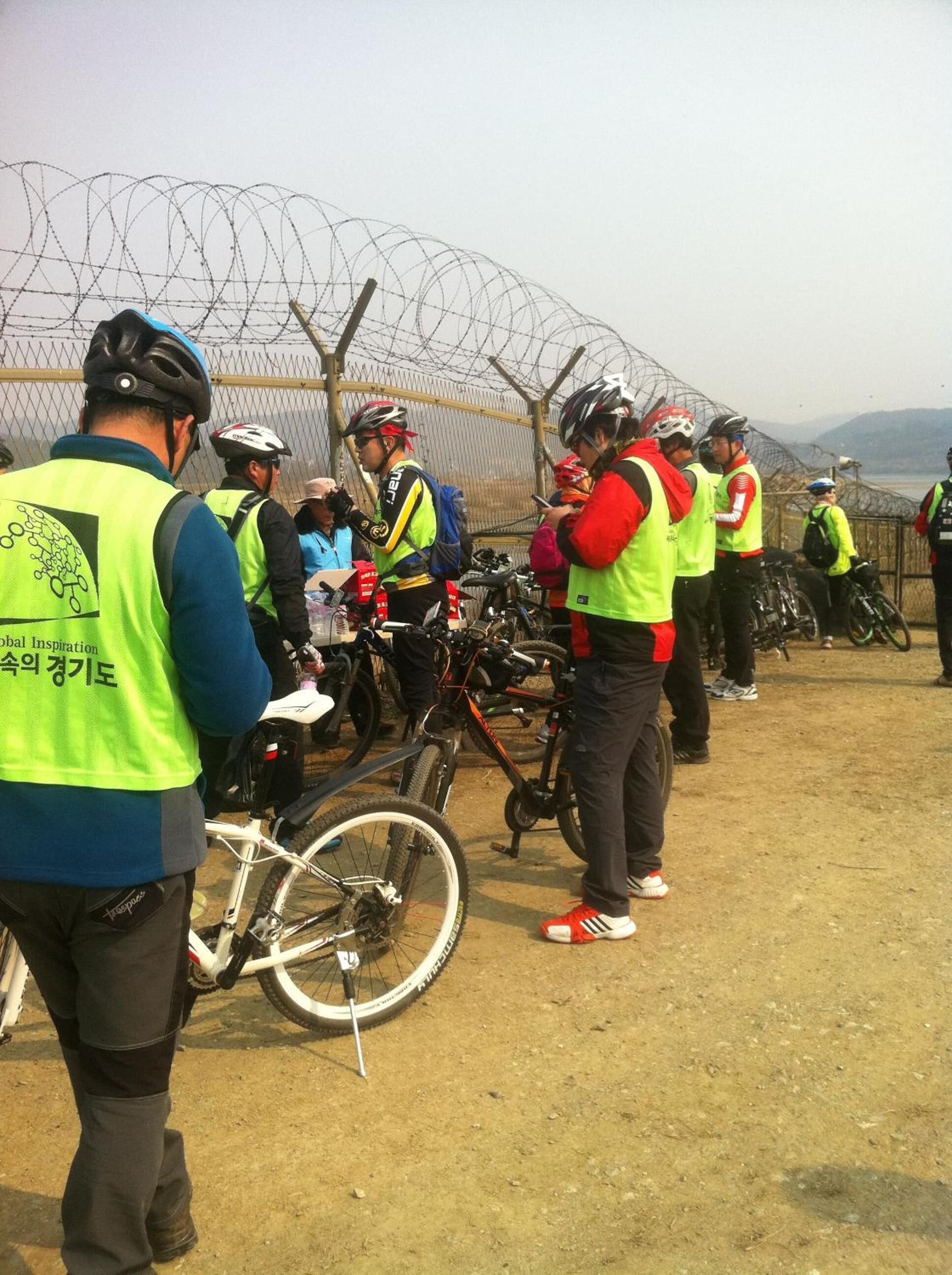 Un grupo de ciclistas reunidos alrededor de sus bicicletas, algunos con chalecos de seguridad, con una cerca de alambre de púas al fondo.
