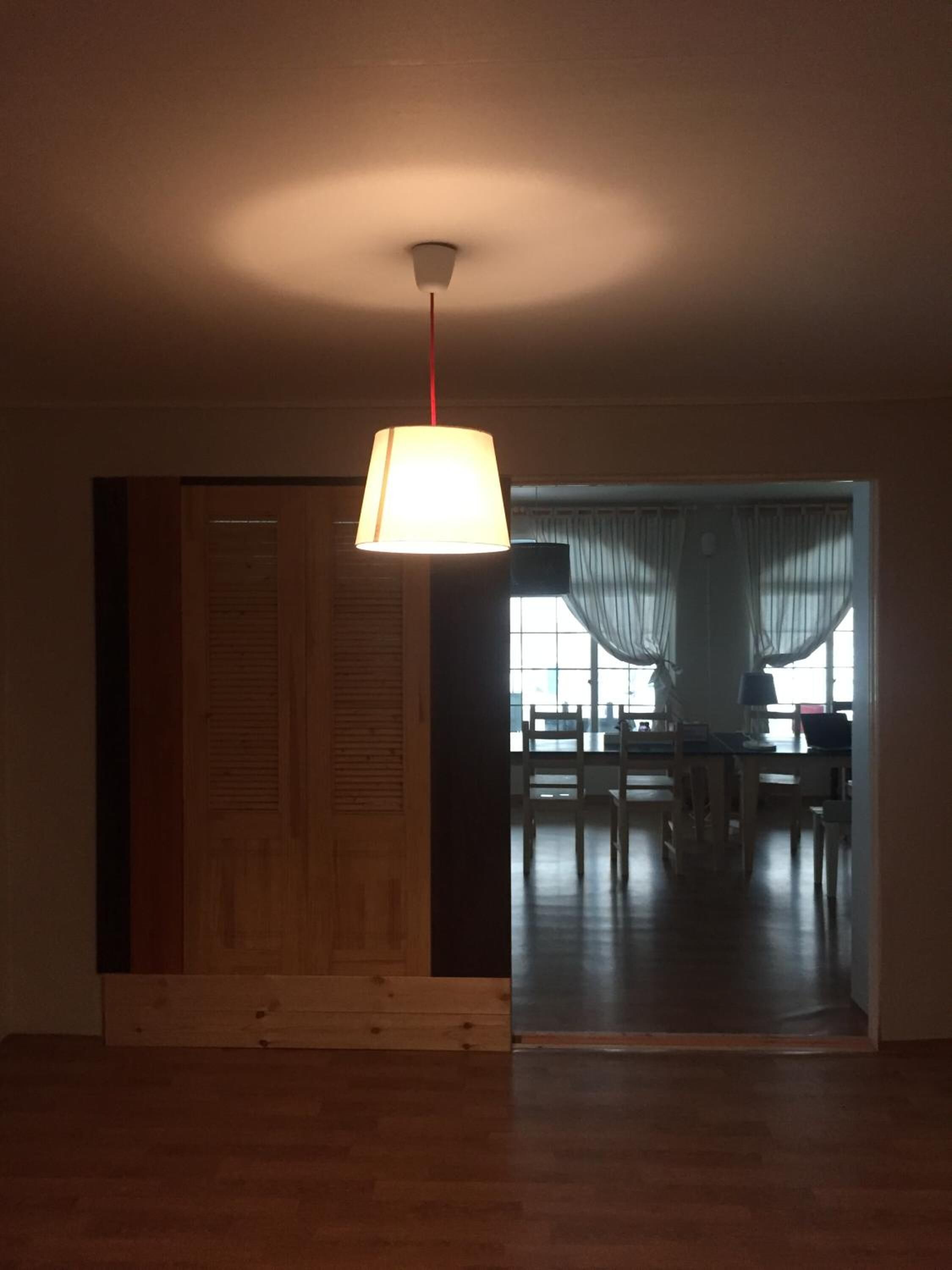 A room with a wooden floor, a hanging lamp, and a view into a dining area with tables and chairs.
