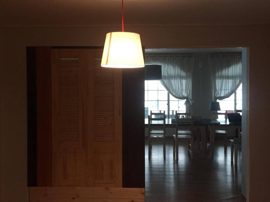 A room with a wooden floor, a hanging lamp, and a view into a dining area with tables and chairs.