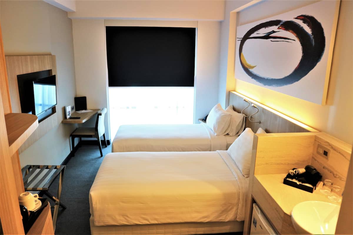 Quarto de hotel com duas camas, uma escrivaninha, televisão, um quadro e banheiro.