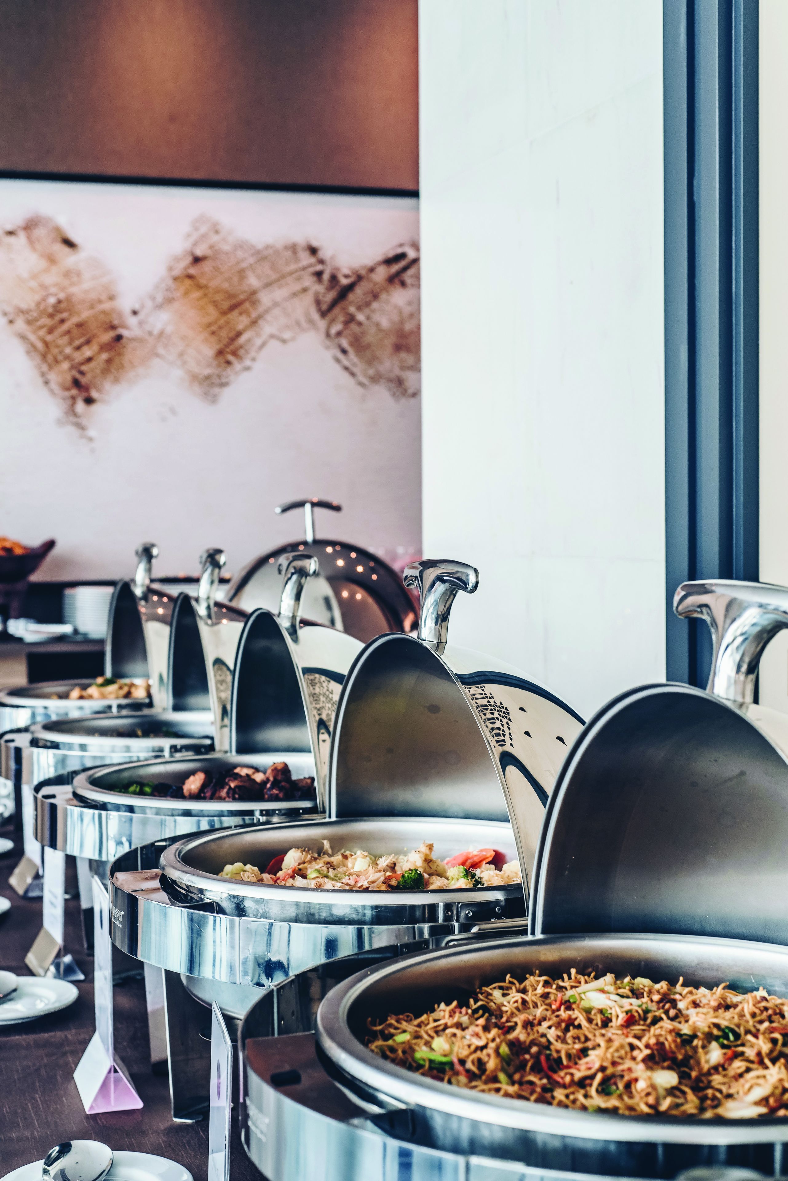 A buffet setup with multiple stainless steel chafing dishes, some containing food, in a modern dining area.