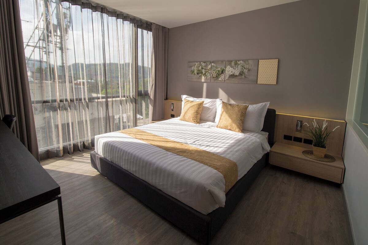 A modern hotel room with a large bed, a bedside table, a nightstand, and a view of the outdoors through sheer curtains.