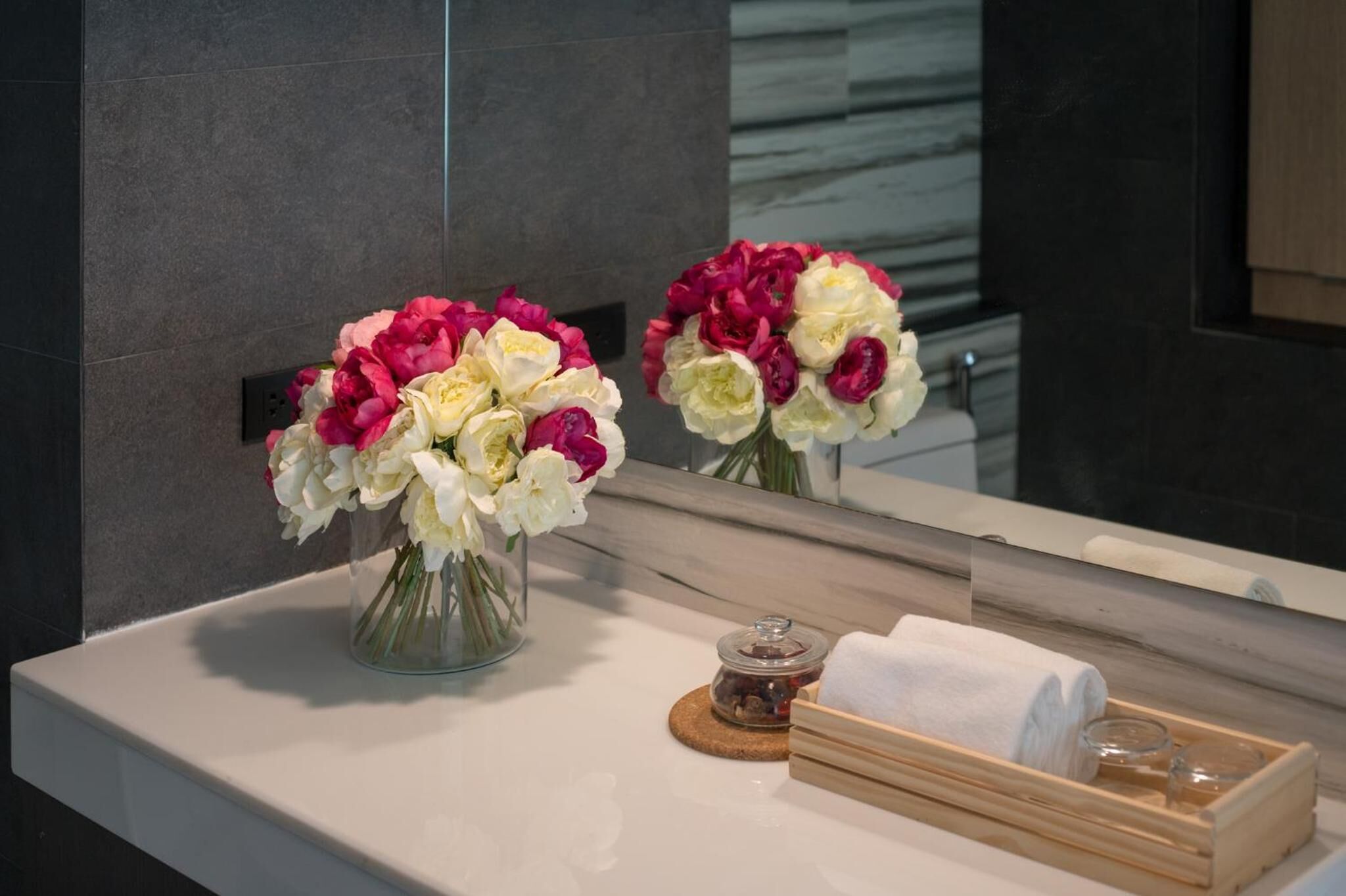 Un bagno con un piano di marmo, due vasi di fiori, un asciugamano e un vassoio di legno con articoli da toilette.