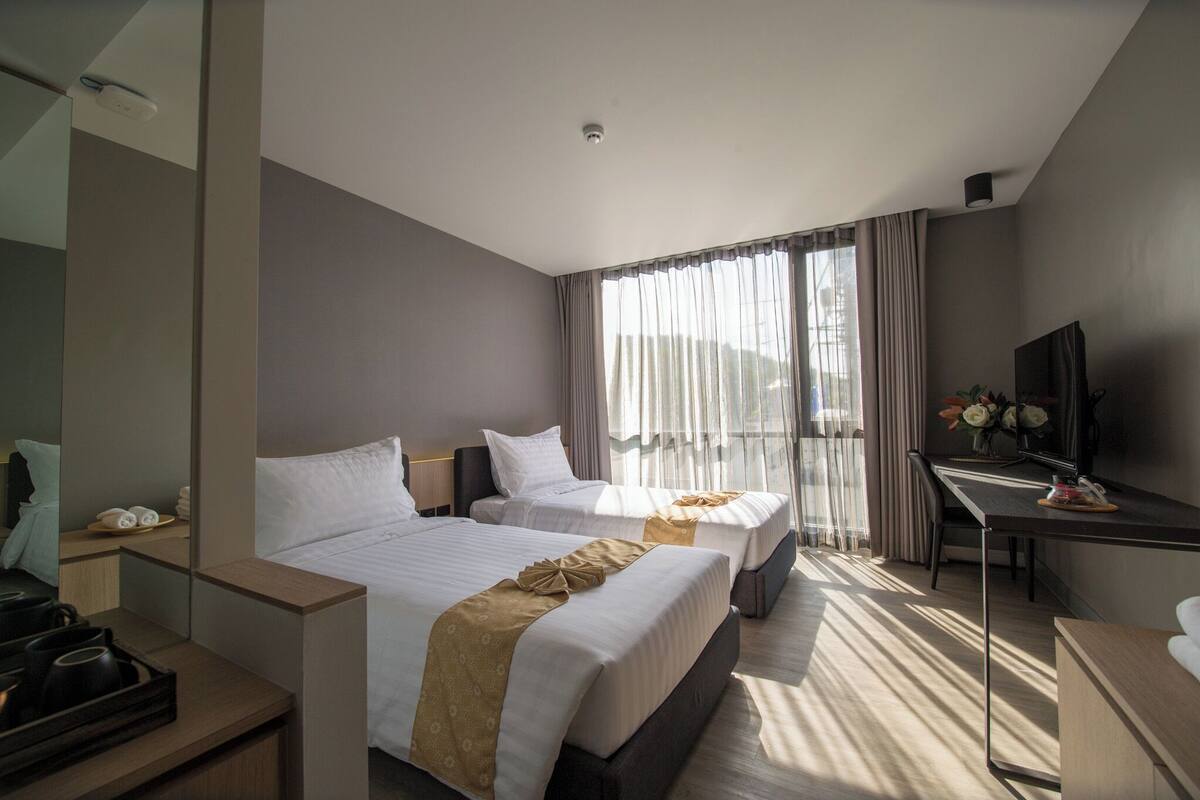 A hotel room with two beds, a desk, a TV, and a large window with curtains.