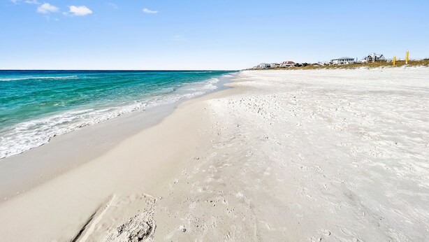 Una playa con agua turquesa cristalina, arena blanca y casas al fondo.