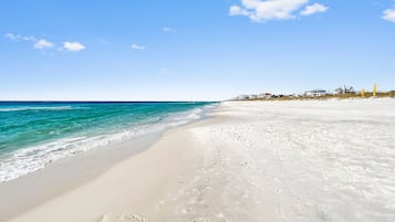 Une plage à l’eau turquoise et limpide, au sable blanc, avec des maisons en arrière-plan.