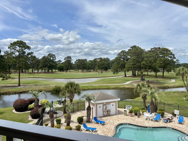A pool area with lounge chairs, a small building, and a golf course in the background. - 4737 Wild Iris Drive Magnolia Place 2 Bedroom Condo by RedAwning (Myrtle Beach)