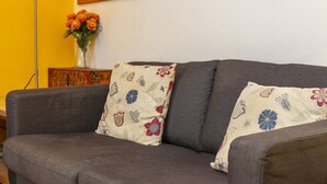 A grey sofa with two decorative pillows featuring floral patterns. A vase with orange flowers is placed on a wooden side table next to the sofa. - A03 - Central 1 Bed Apartment (Lagos)