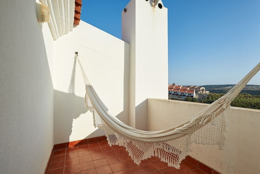 Eine weiße Hängematte auf einem gefliesten Balkon mit freiem Blick auf Gebäude und Grünanlagen im Hintergrund.