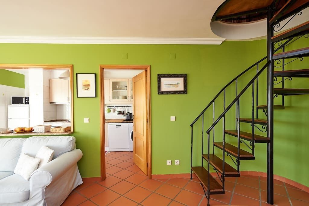 A living room with a white sofa, a staircase with a metal railing, and a kitchen visible through an open door.