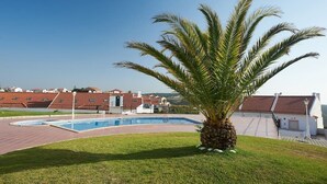 A swimming pool with a palm tree in the foreground, surrounded by a grassy area and buildings with red-tiled roofs in the background. - CORAL VILLA, a charming landscape close to the beach, best choice for families (Carvoeira)
