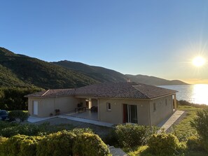 Une maison de plain-pied avec un toit en tuiles, une terrasse dotée de mobilier d’extérieur et une vue sur la mer et les montagnes en arrière-plan.