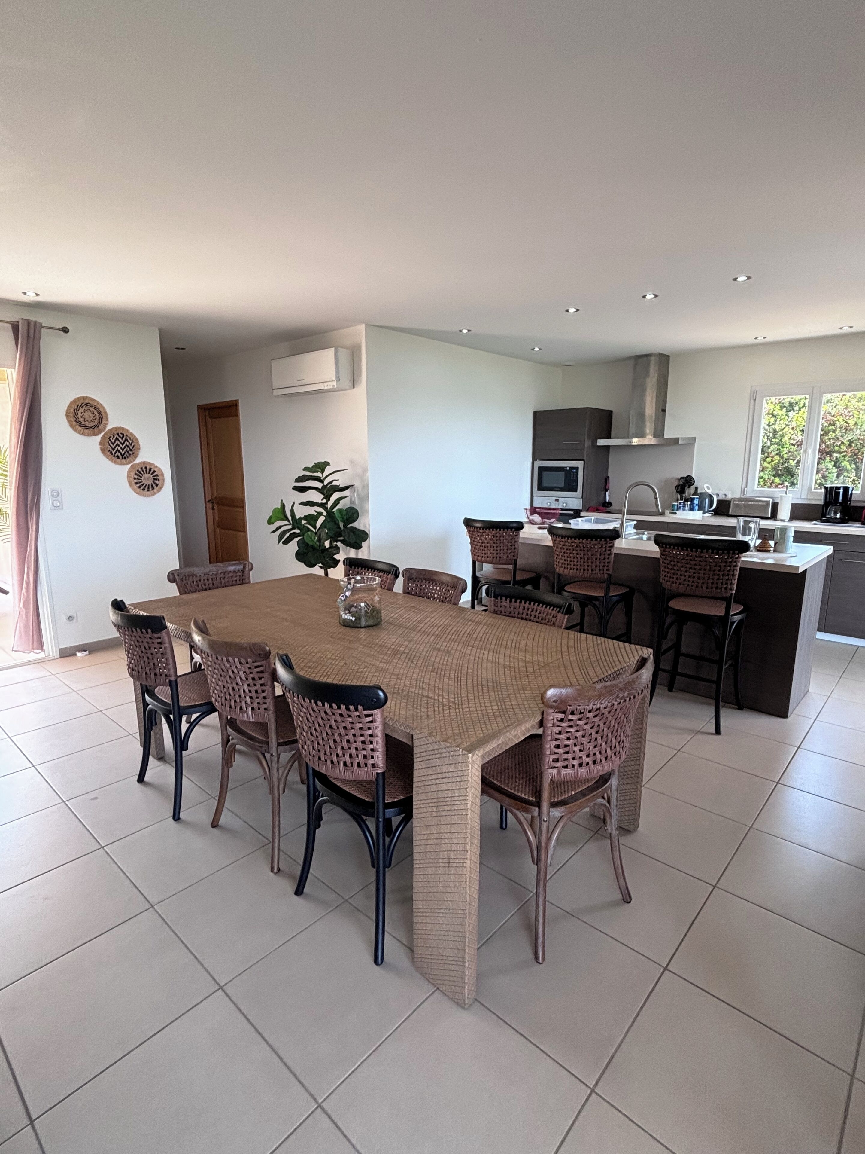 A modern kitchen with a central island, a dining area with wicker chairs, and a large window providing natural light.