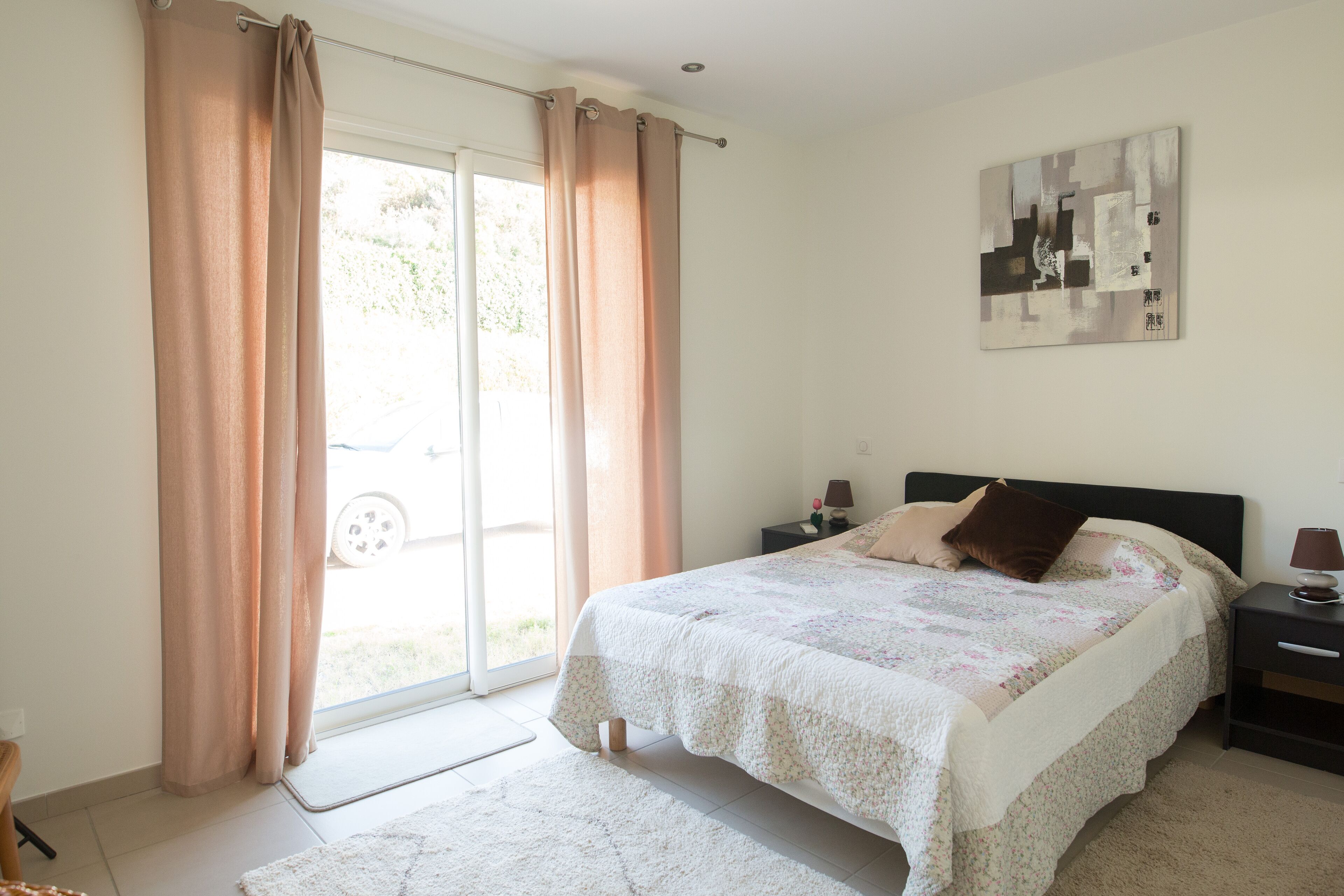 A bedroom with a bed, nightstands, a lamp, a painting, and a sliding glass door.