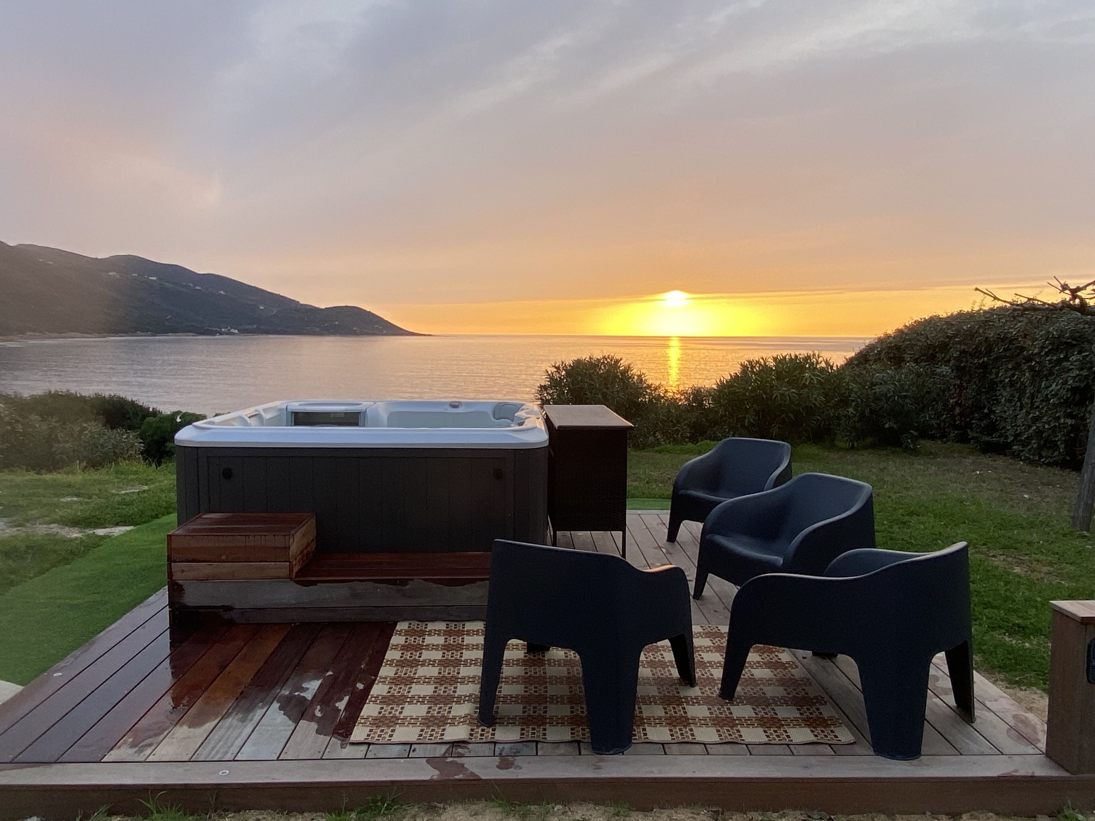 Boblebad på en træterrasse med stole, med udsigt over vand ved solnedgang.