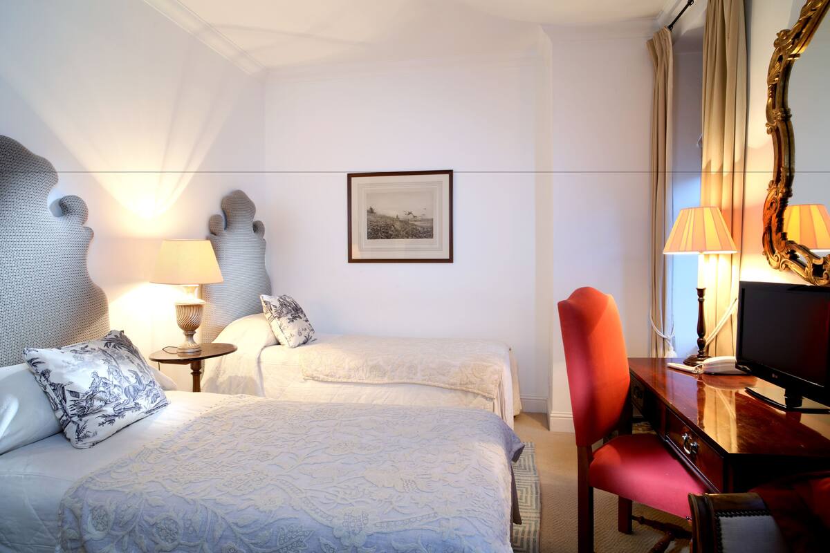 Hotellihuone, jossa on kaksi sänkyä, työpöytä, jossa on tietokone, tuoli, lamppu ja seinällä oleva kehystetty kuva.