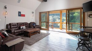 A spacious living room with a leather sofa, a coffee table, and a dining area with a table and chairs. Large sliding glass doors lead to a balcony with a view of trees.