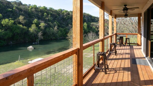 A wooden deck with a railing overlooking a river, a table, and chairs.