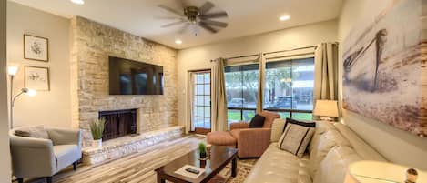 A living room with a stone fireplace, a ceiling fan, and a large window with curtains.