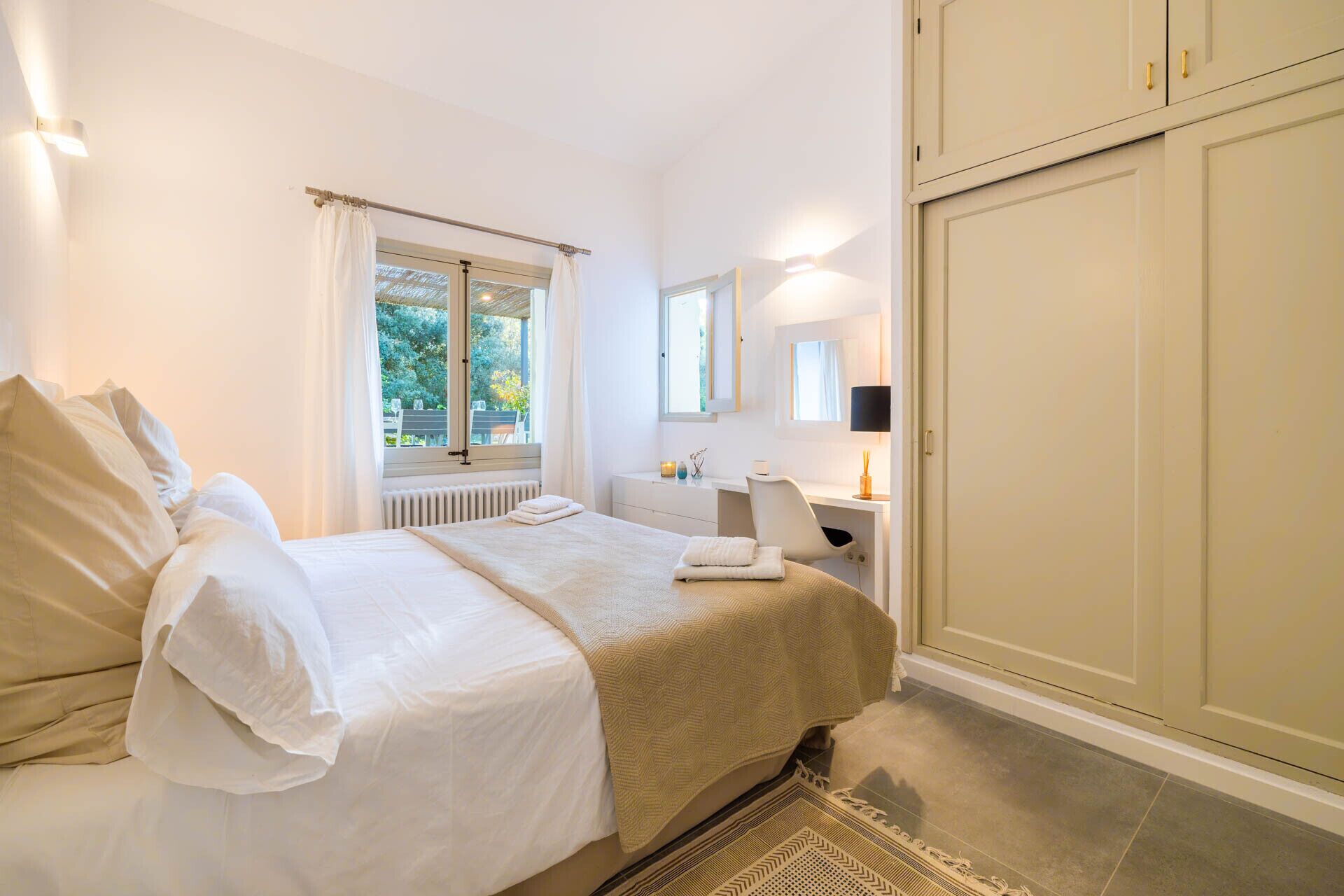 A bedroom with a bed, a window, a bedside table, a mirror, and a wardrobe.