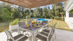 Un patio exterior con una mesa preparada para el desayuno, rodeado de sillas, con una piscina y abundante follaje de fondo.