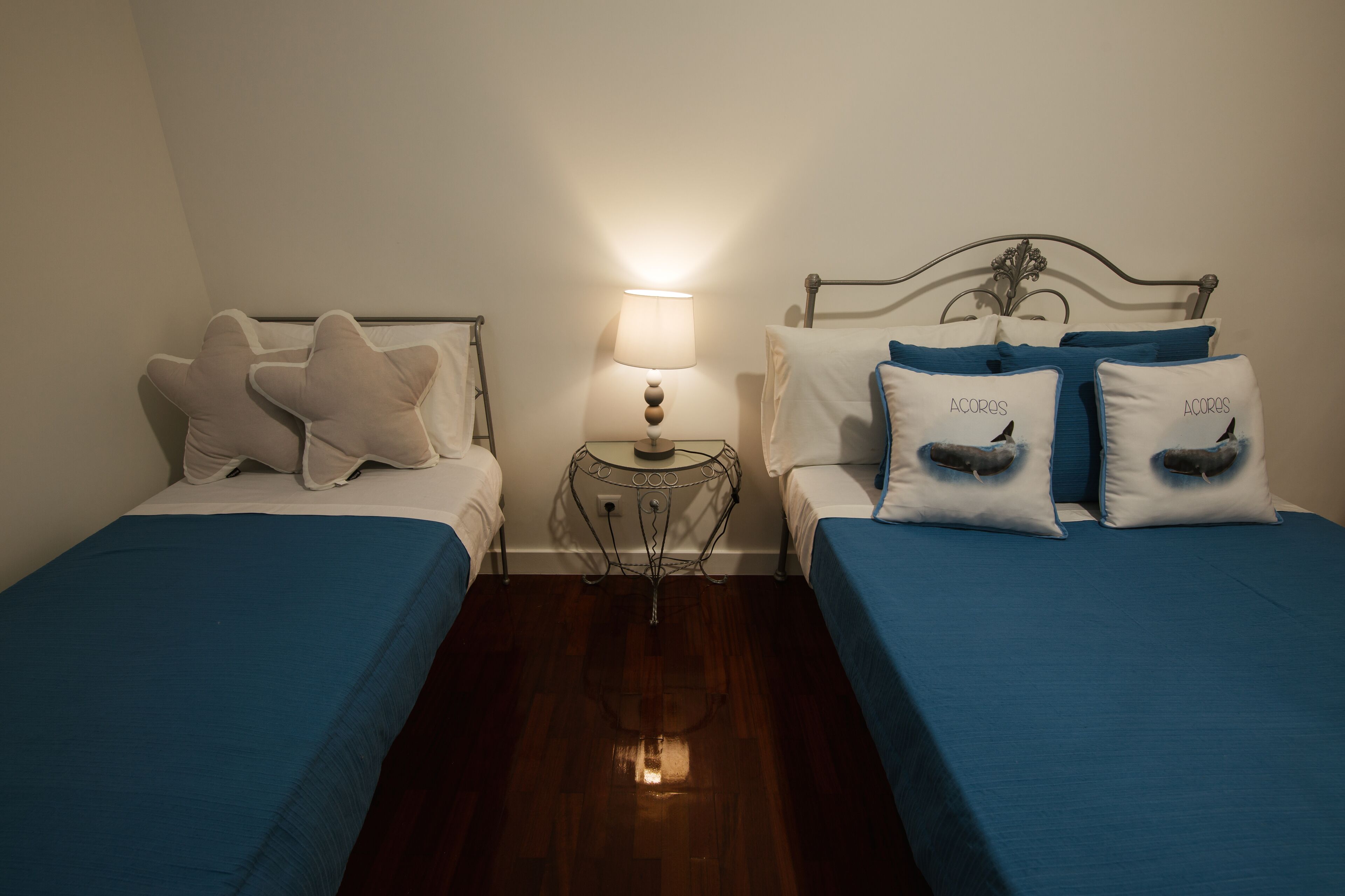 A hotel room with two beds, each with blue bedding and decorative pillows, a bedside table with a lamp, and a small round mirror.