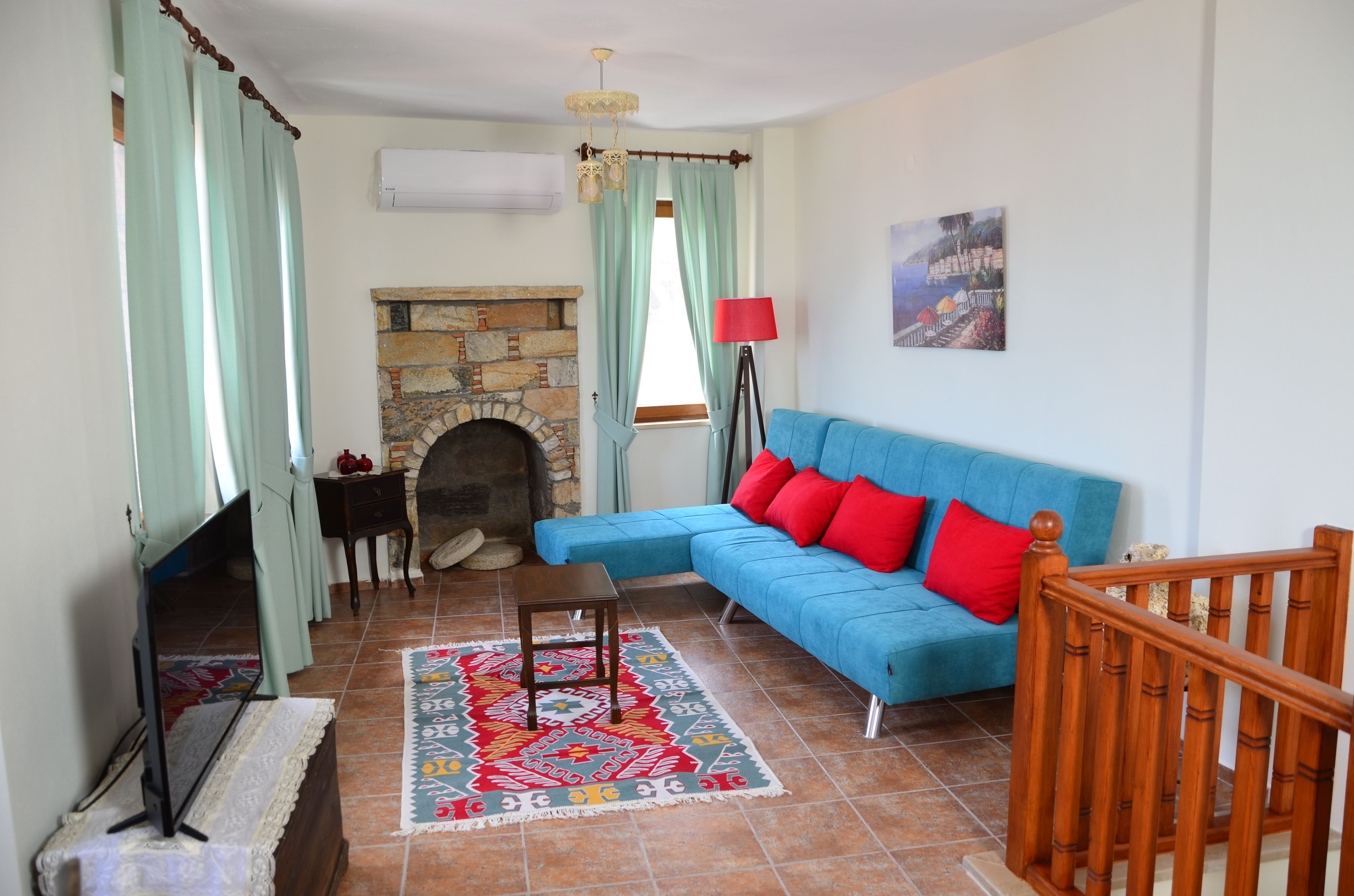 Una sala de estar con chimenea de piedra, un sofá azul con cojines rojos, un tapete estampado y una barandilla de madera para las escaleras.