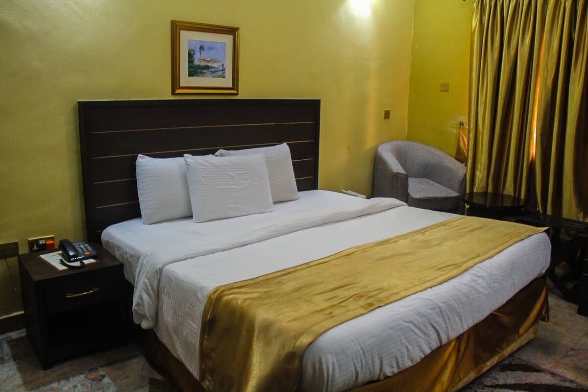 A hotel room with a large bed, a bedside table with a phone, a chair, and a curtain.