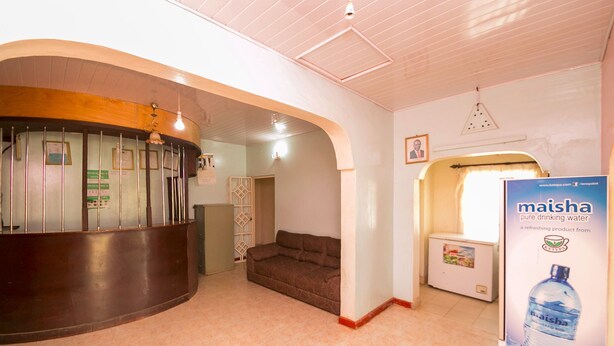 A reception area with a curved reception desk, a sofa, a refrigerator, and a poster advertising Maisha water. - Legacy 2014 Hotel (Bondo)