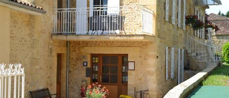 A stone building with a wooden door, a balcony with white railings, and a flower planter in front.