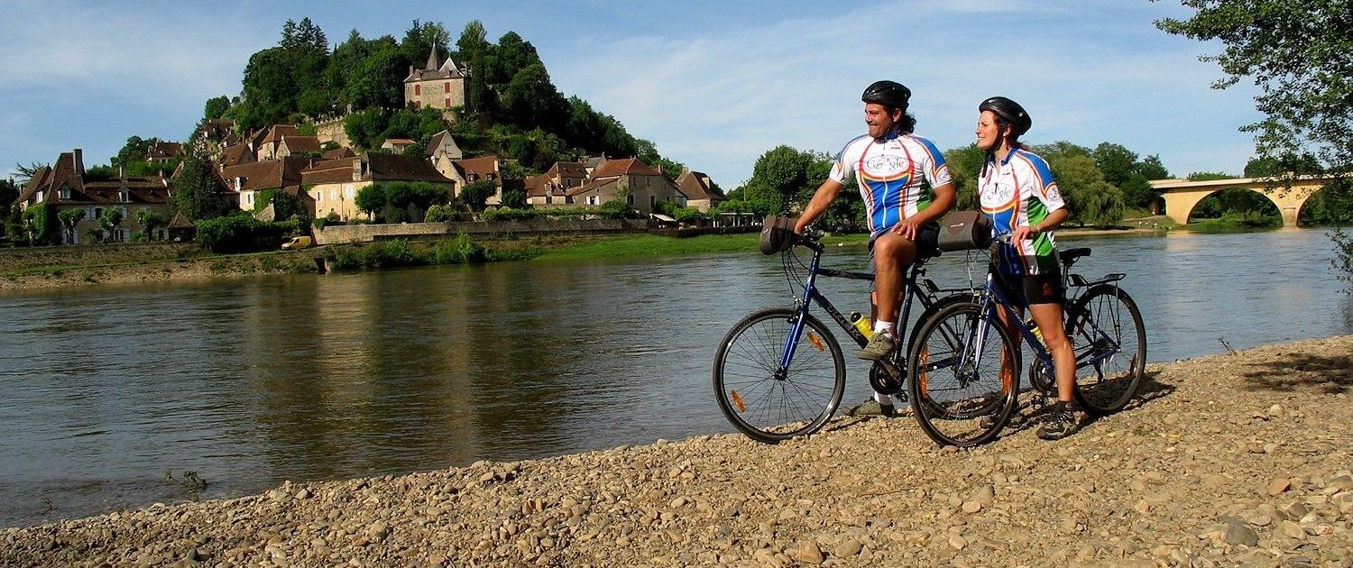 Zwei Radfahrer an einem Fluss, im Hintergrund ein historisches Gebäude.