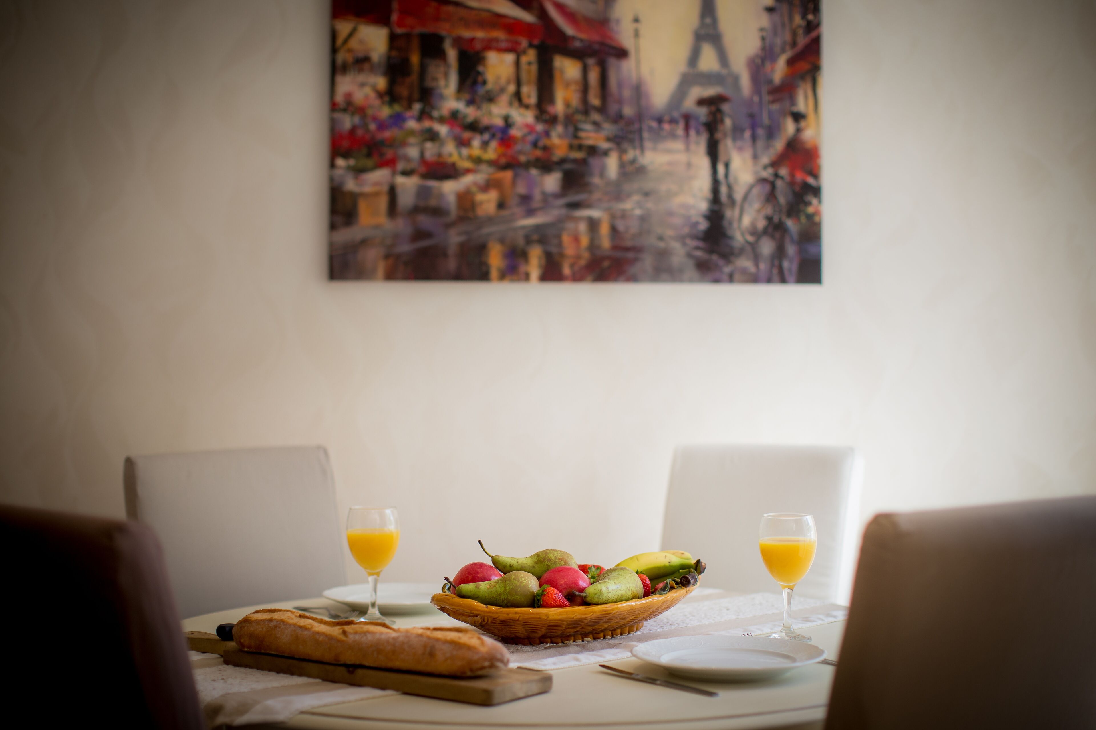 Una mesa de comedor con una cesta de fruta, dos vasos de zumo de naranja, una baguette y un cuadro de una escena callejera.
