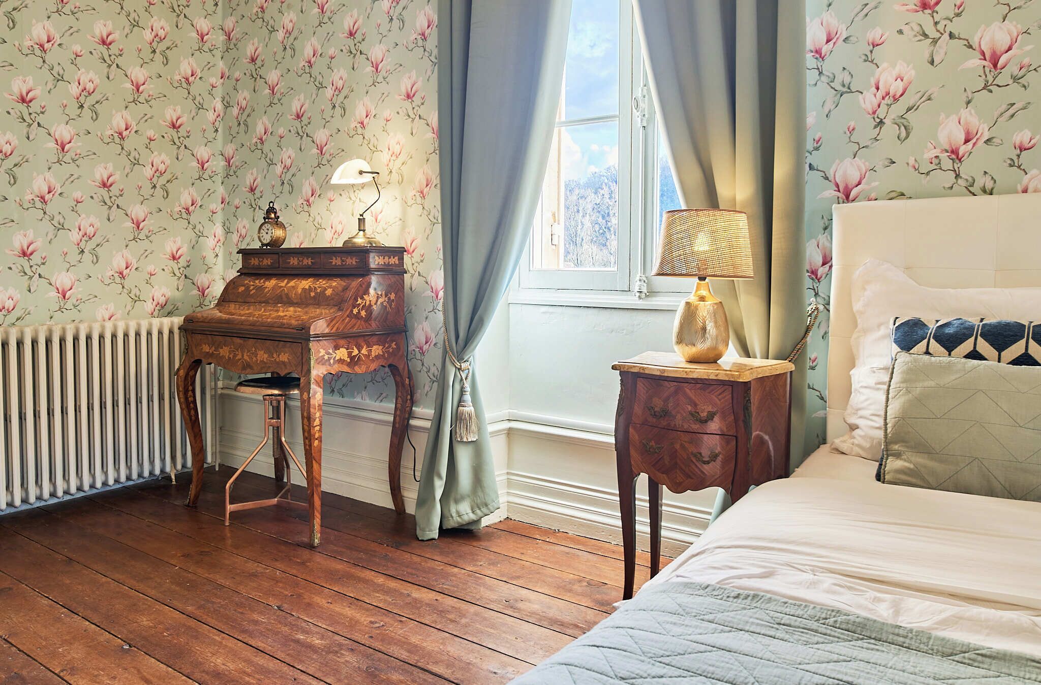 A room with floral wallpaper, a wooden piano, a radiator, a window with curtains, a bedside table with a lamp, and a bed with pillows.