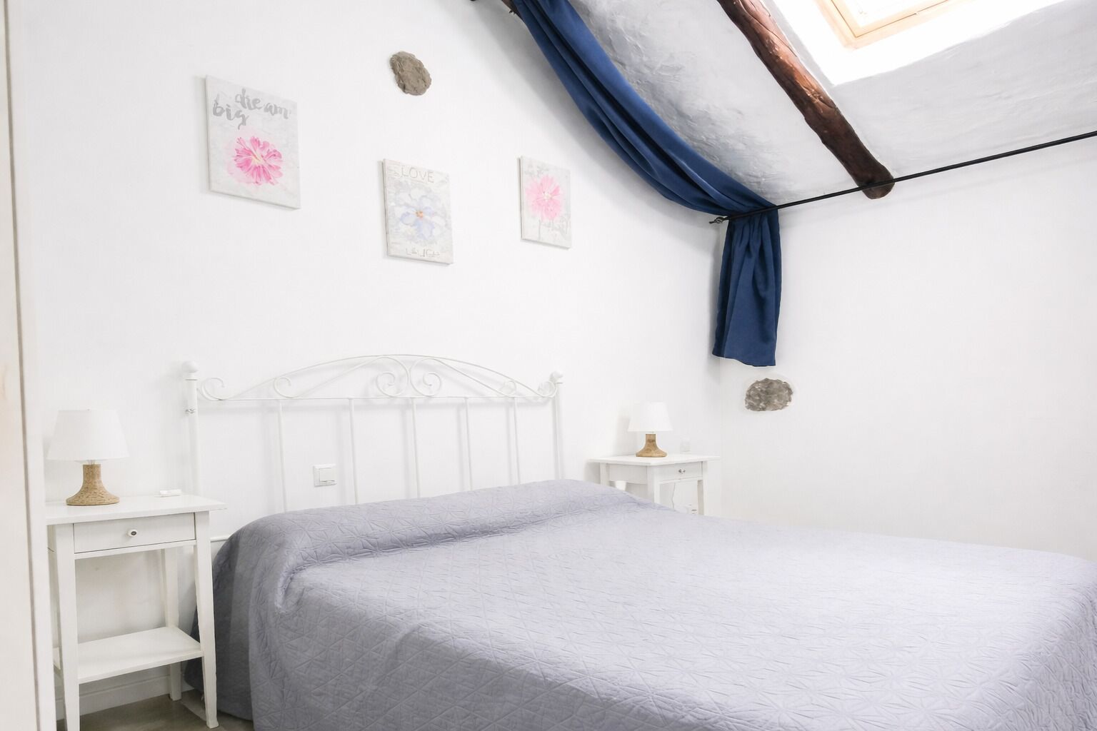 Un dormitorio con una cama blanca, una mesita de noche con lámpara, una pequeña mesa con lámpara y decoración en la pared.