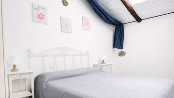 Un dormitorio con una cama blanca, una mesita de noche con lámpara, una pequeña mesa con lámpara y decoración en la pared.