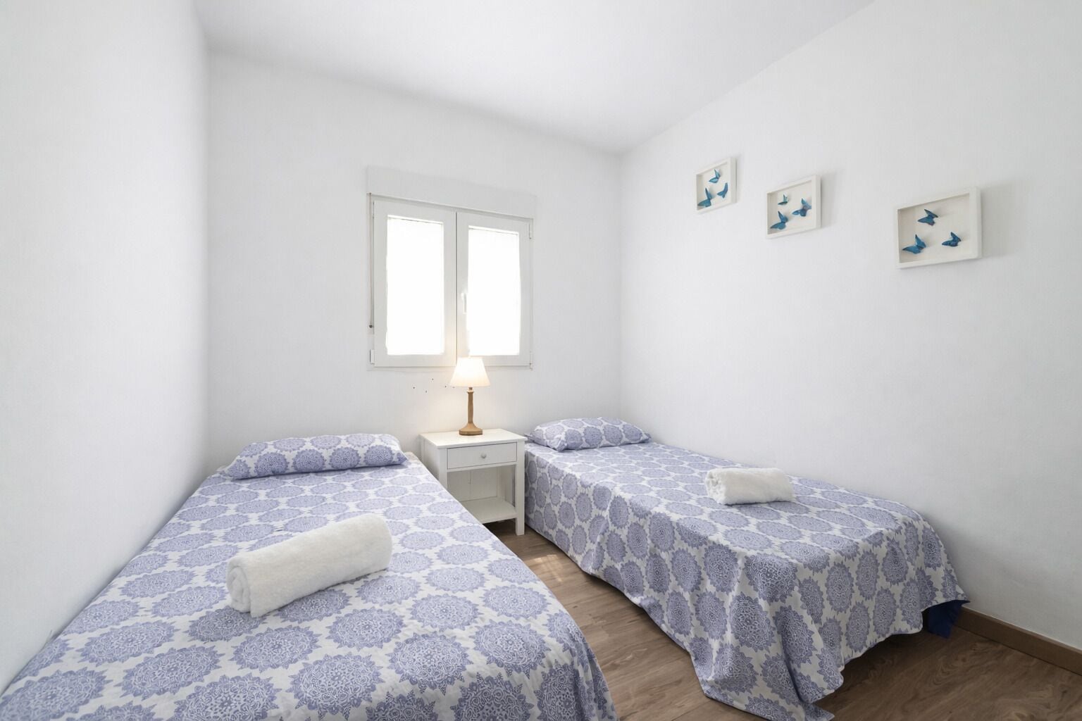 Une petite chambre propre, avec deux lits simples, une table de chevet, une lampe et une fenêtre.