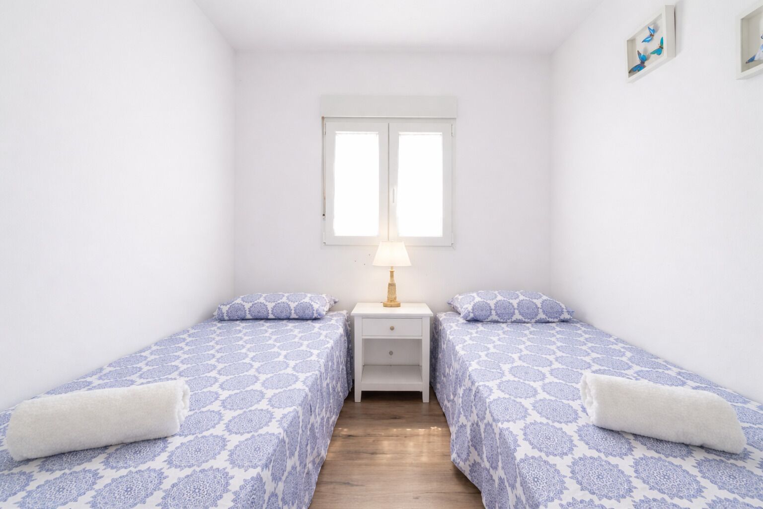 Une petite chambre propre, avec deux lits simples, une table de chevet avec une lampe et une fenêtre avec des stores.