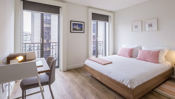 A modern bedroom with a large bed, a desk with a chair, and large windows offering a city view.