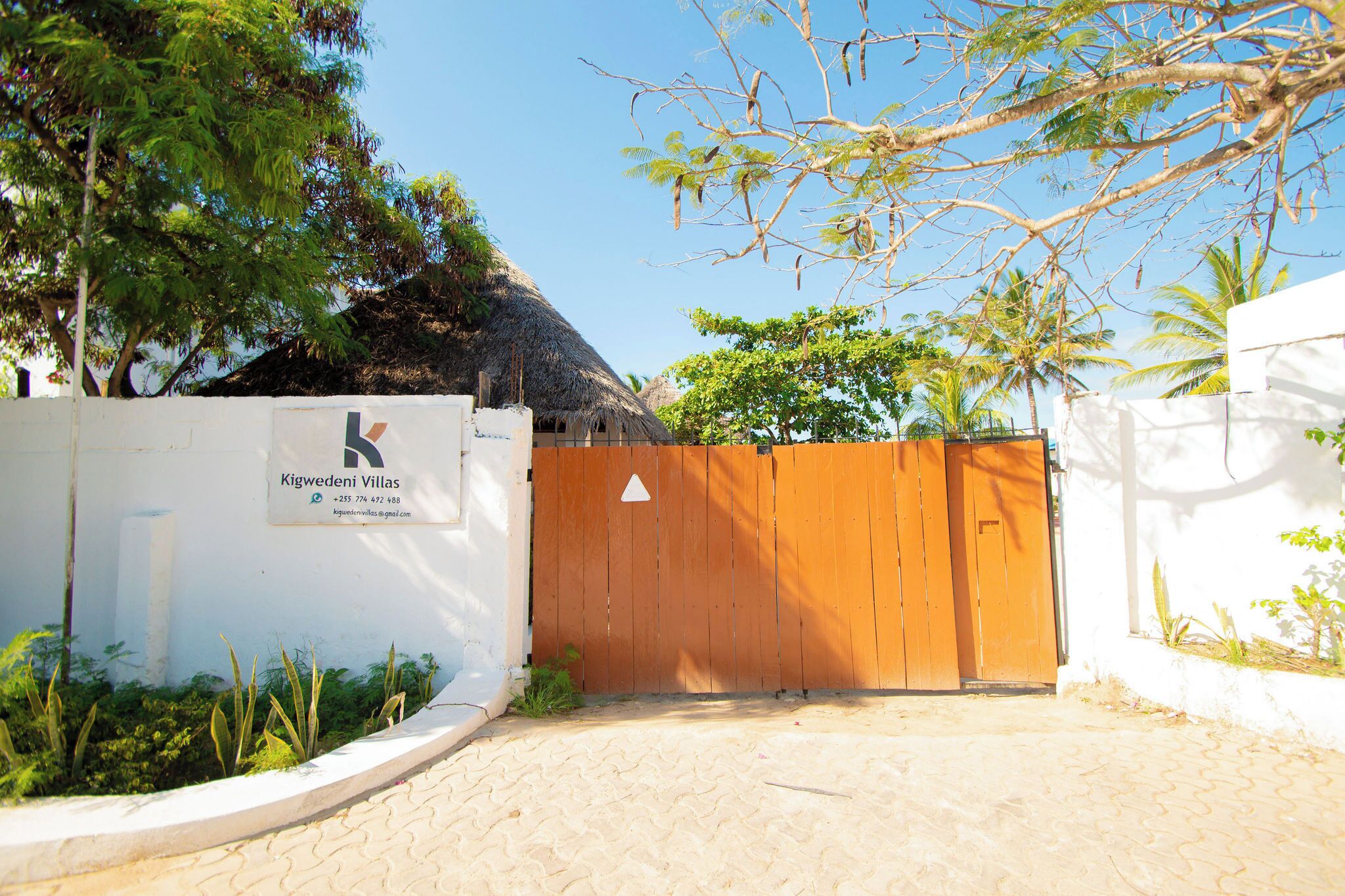 A gated entrance with a sign for 'Kigwedeni Villas,' a thatched-roof structure, and surrounding greenery.