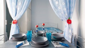 A neatly set dining table with blue curtains, a bed with a blue and white patterned bedspread, and a red flower decoration. - Wavedance Accommodation (Salamis)