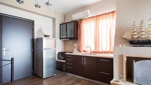 A compact kitchen with a refrigerator, microwave, and a small sink. There is a window with curtains and a decorative ship model above the fireplace. - Wavedance Accommodation (Salamis)