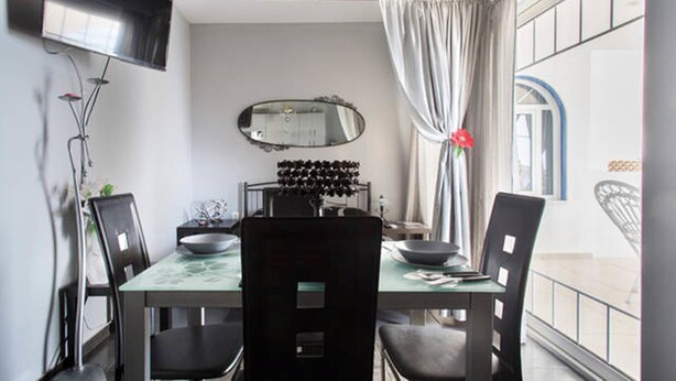 A dining area with a table, chairs, a flat-screen TV mounted on the wall, a mirror, and a window with curtains. - Wavedance Accommodation (Salamis)