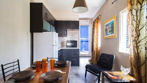 A modern kitchen with dark cabinets, a white refrigerator, and a microwave. There is a dining table with plates and glasses, and a chair. A window with curtains is visible, and a painting hangs on the wall. - Wavedance Accommodation (Salamis)