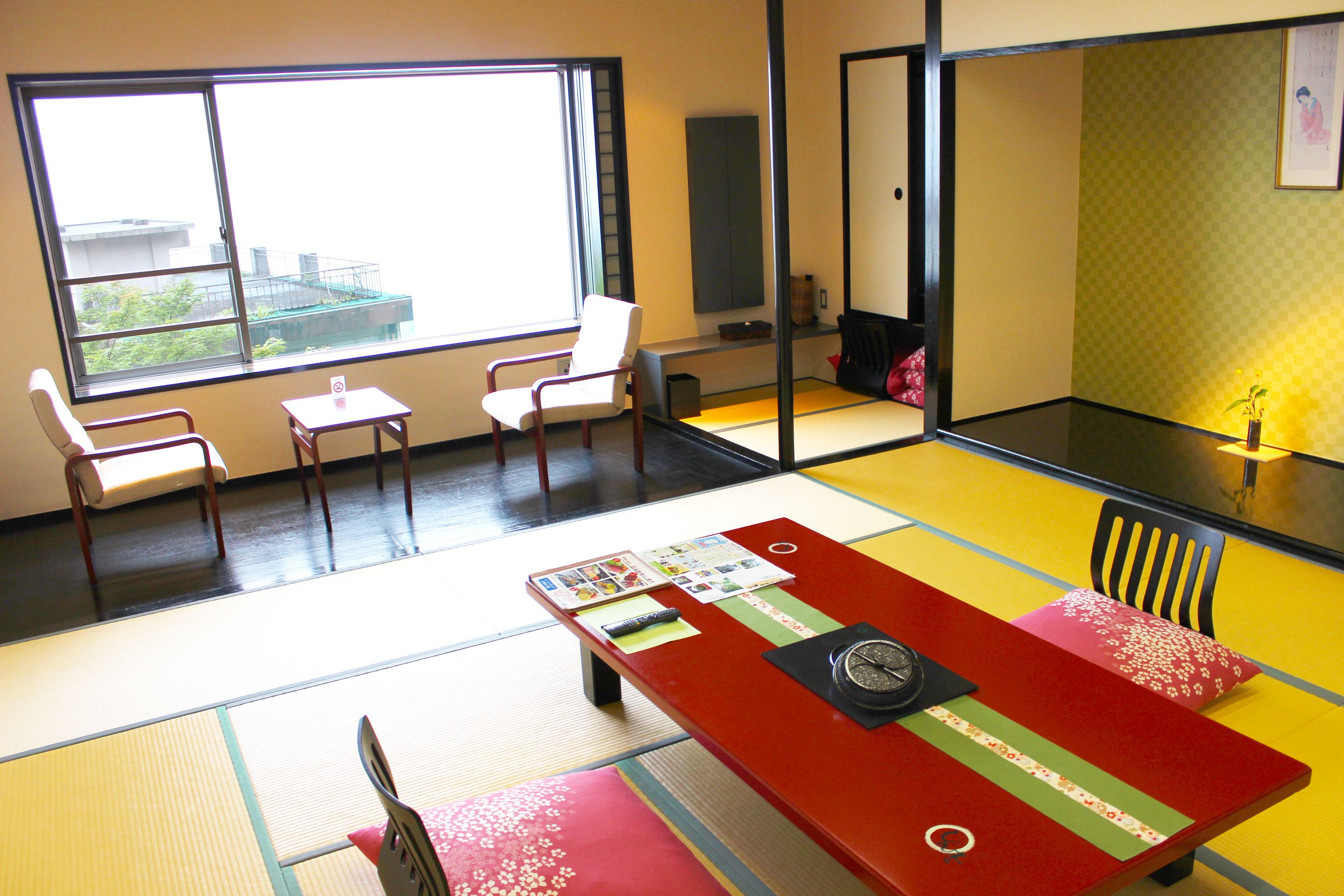 A room with tatami flooring, a low table with a clock, and a view of buildings outside a large window.