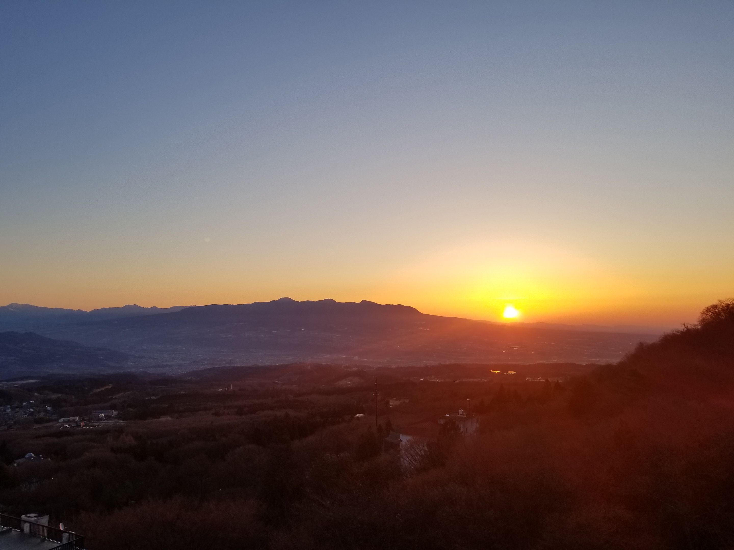 Un coucher de soleil sur un paysage montagneux, avec vue sur une vallée et quelques bâtiments.